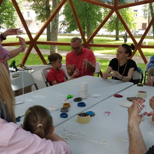 Prie stalo sėdintis žmonės rankose pina vilnonius siūlus.