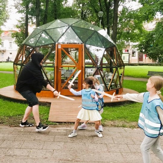 Vaikai mėlynomis  atšvaitinėmis liemenėmis ir vyras su juodu džemperiu kovoja tarpusavyje su mediniais kalavijais. 