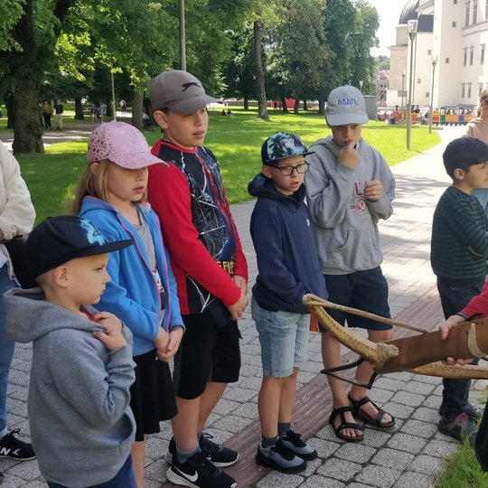 Viduramžiškai apsirengęs vaikinas susirinkusiems vaikams rodo ir pasakoja apie arbaletą.