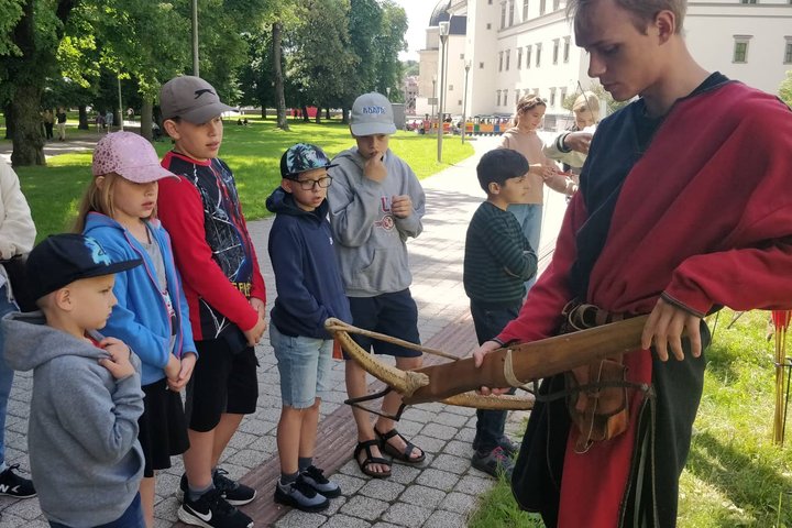 Viduramžiškai apsirengęs vaikinas susirinkusiems vaikams rodo ir pasakoja apie arbaletą.