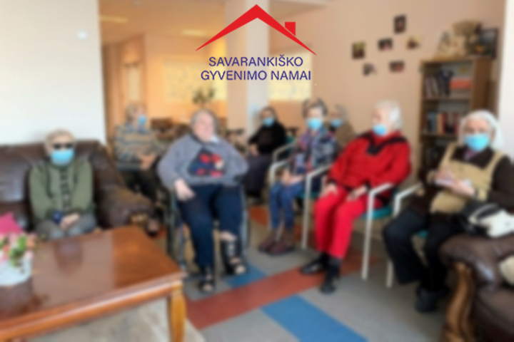 Nemenčinės „Savarankiško gyvenimo namų“ gyventojai kambaryje. Nuotraukos centre, viršuje Nemenčinės „Savarankiško gyvenimo namų“ logotipas.