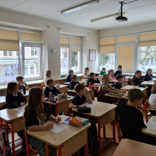 Trečiokėliai sėdi klasės suoluose ir klausosi edukatorės vedamos pamokos.