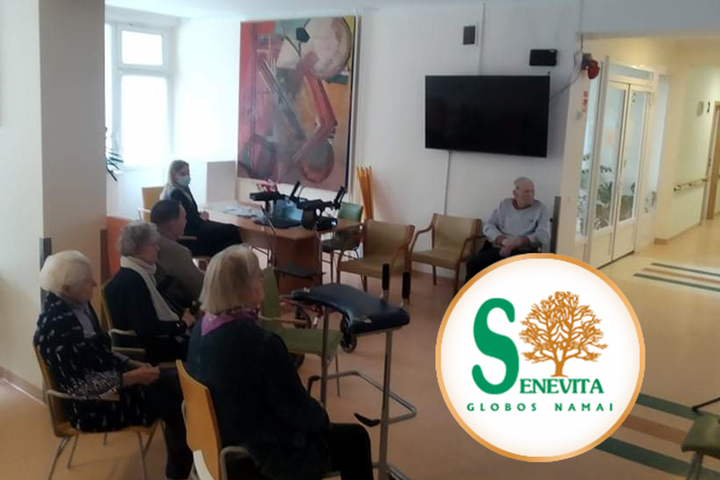 Senjorai, sėdėdami salėje stebi ekraną. Nuotraukos kampe „Senevita“ logotipas.