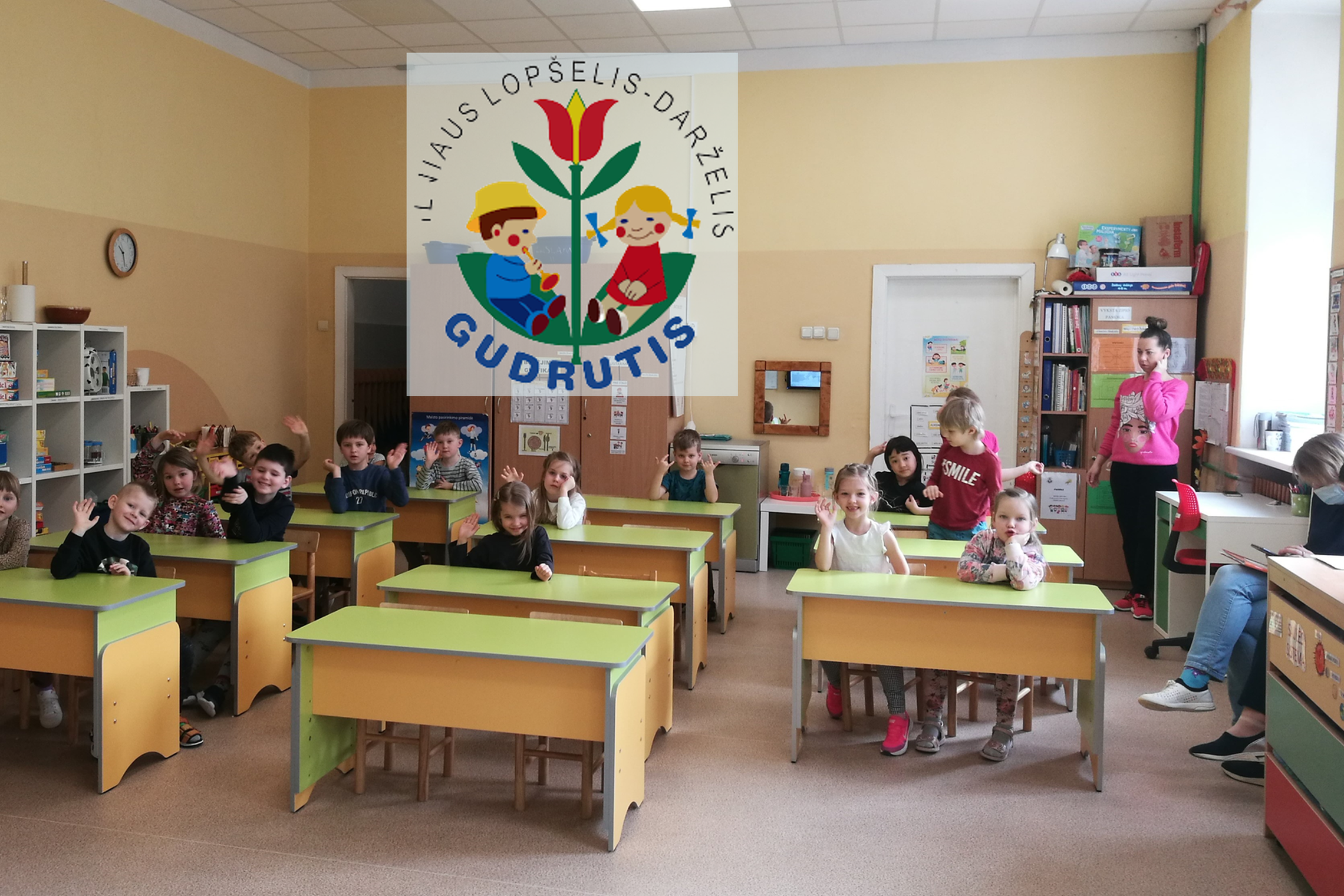 Darželinukai klasėje, sėdi prie suolų. Nuotraukos Viršuje, centre lopšelio – darželio "Gudrutis" logotipas.