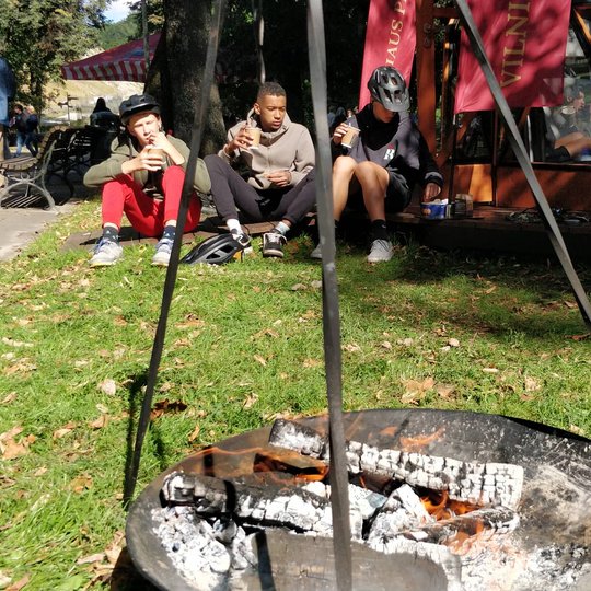 Po ugnies įskėlimo, galime džiaugtis ir rezultatu - mėtų arbata. Trys vaikinai geria arbatą prie Vilniaus pilių kupolo. Priekyje dar rusenantis laužas.
