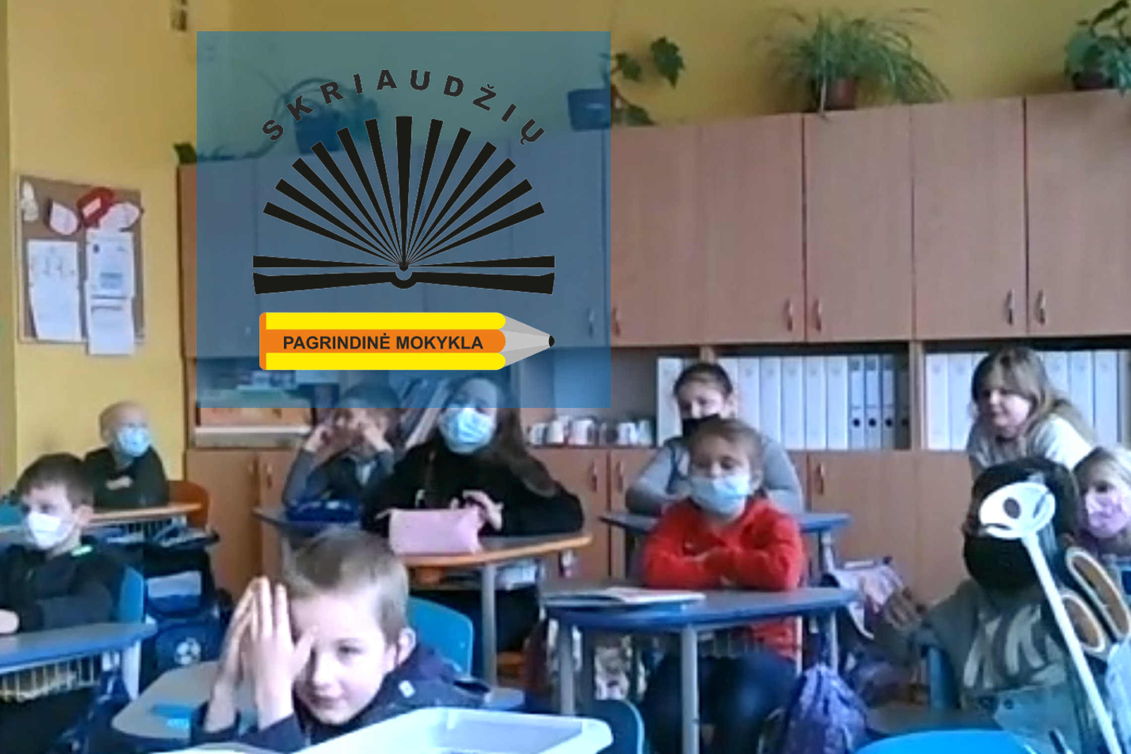 Mokiniai sėdi prie suolų klasėje. Nuotraukos priekyje Skriaudžių pagrindinės mokyklos logotipas.