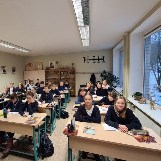 Klasė, pilna mokinių. Mokytoja stovi priekyje. Vaikai ruošiasi gaminti savo herbus.