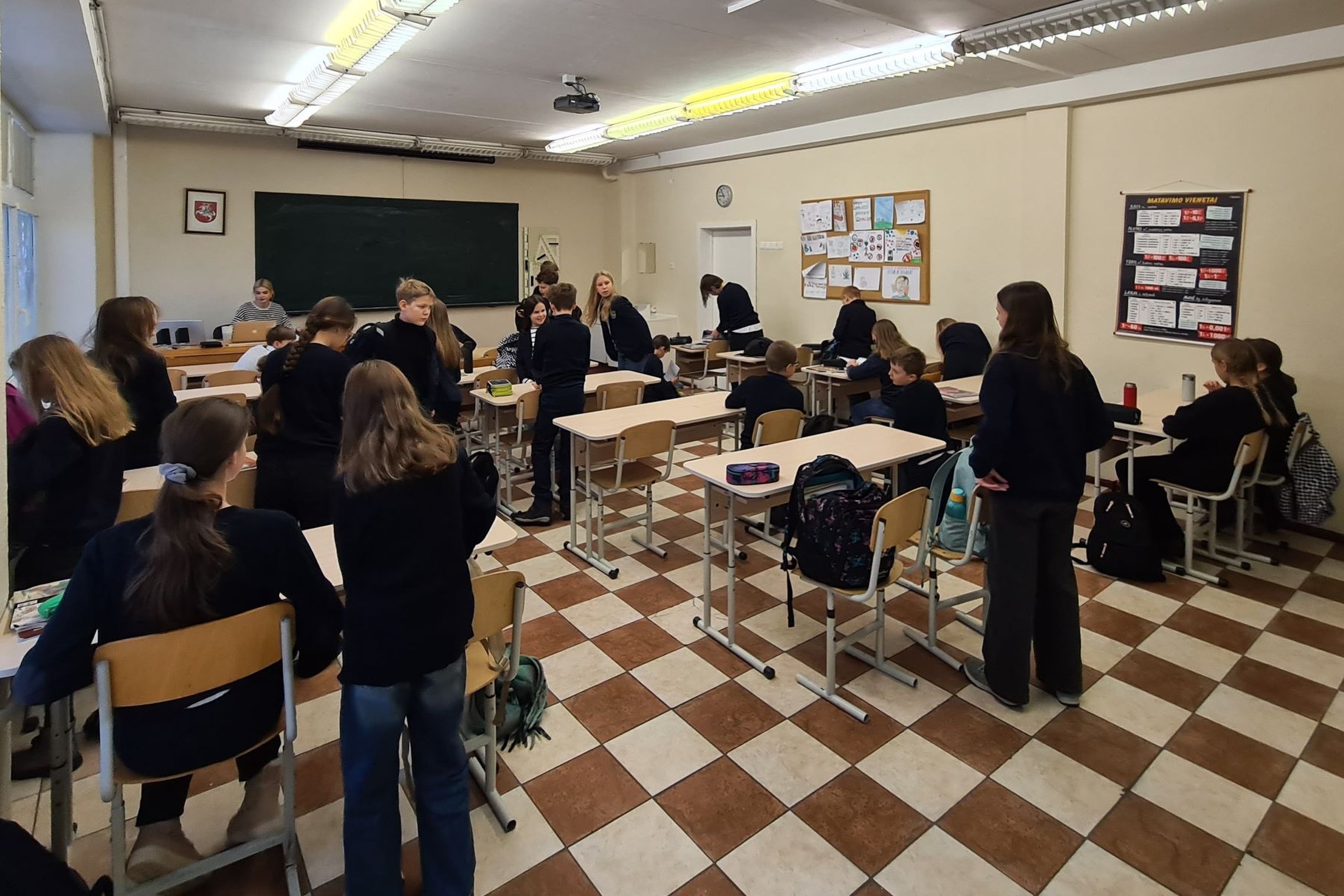 Mokiniai klasėje stovi prie suolų ir ruošiasi pamokai.