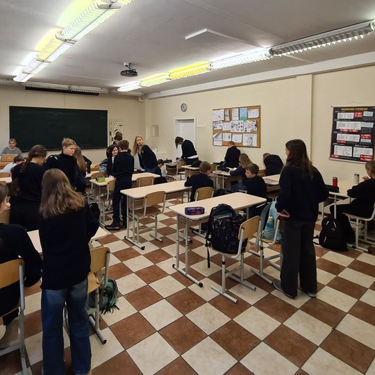 Mokiniai klasėje stovi prie suolų ir ruošiasi pamokai.