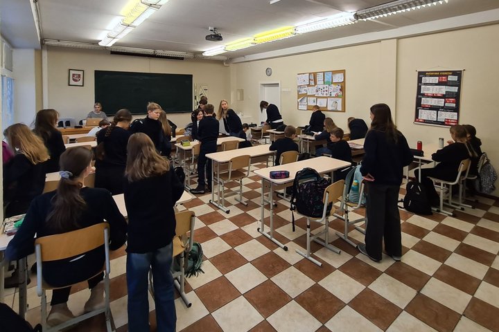 Mokiniai klasėje stovi prie suolų ir ruošiasi pamokai.