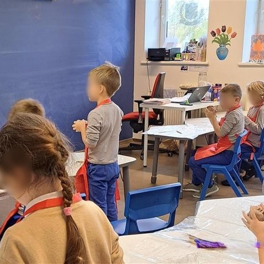 Mokytoja su prijuoste stovi prie juodos lentos ir rodo vaikams, kaip formuoti molio dirbinį. Pradinukai su raudonomis prijuostėmis atidžiai stebi ir laiko molio gabalėlius rankose. Klasėje matyti stalai, kompiuteris, langai su dekoracijomis ir vaikų kūryb