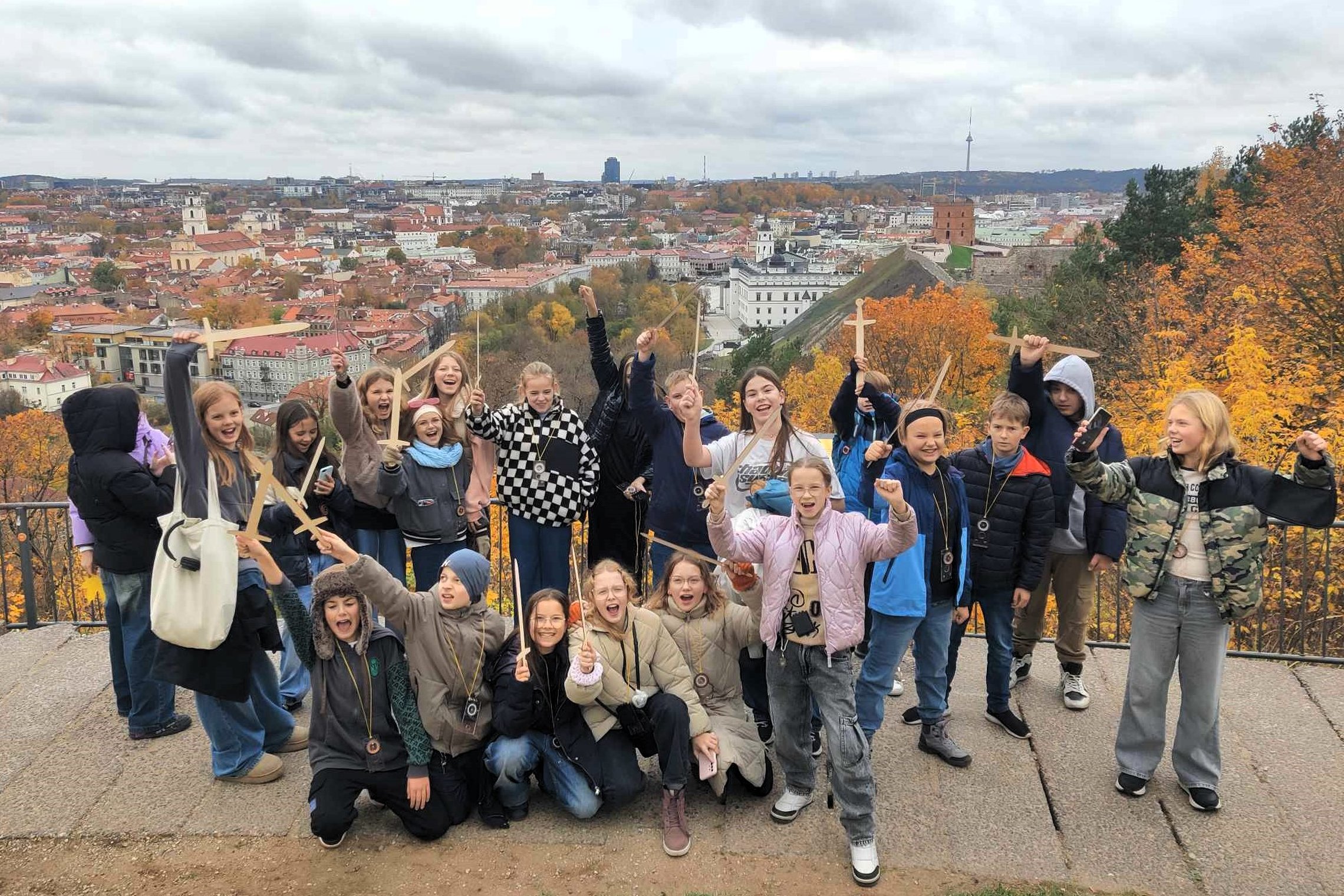 Vaikų būrys pozuoja nuotraukai, visi rankose laiko medinius kardus. Už jų kalno papėdėje matosi senamiesčio pastatų stogai, Gedimino pilis, Valdovų rūmai, rudenio spalvomis pasipuošę medžiai. 