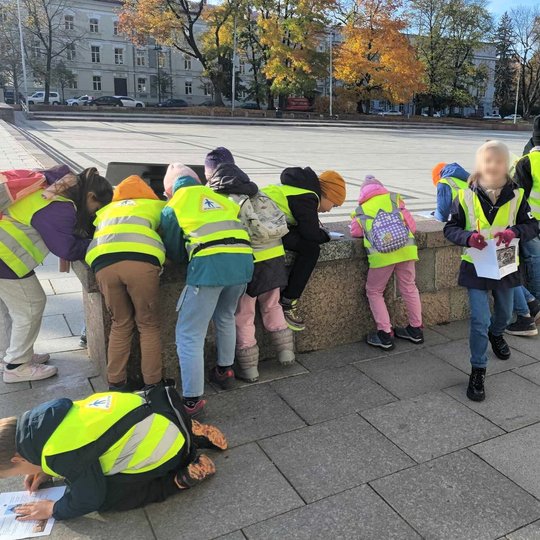 Nuotraukoje galime matyti vaikus, dėvinčius geltonas liemenes. Vaikai stebi Kunigaikščio Gedimino paminklą. taip pat fone galime matyti dalį Gedimino pilies sienos bei šalimais kalno esančio mūrinio namo kampą.