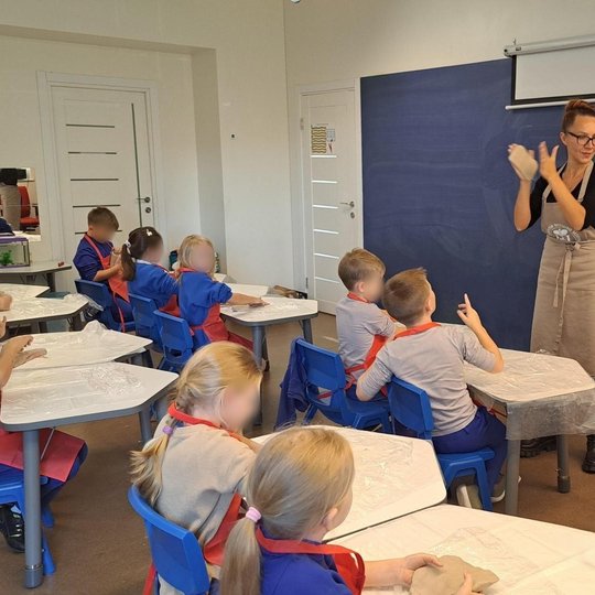 Vaizduojama pradinukų klasė, vaikai, apsirengę raudonomis prijuostėmis, minko molį. Priešais klasę stovi dėtytoja, kuri iškelui molį demontruoja kaip lipdyti. Kabinetas šviesus su mėlynais akcentais.