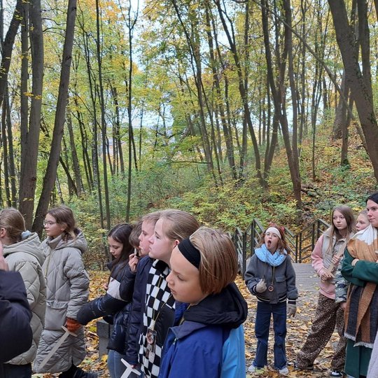 Nuotrauka: vienuolika vaikų stovi ir klausosi ką pasakoja moteris priekį jų. Dvi moterys stovi gale. Panašu, kad vyksta klasės ekskursija miške, kur aplinkui daug geltonų medžių. Žemė nuklota geltonais lapais. Fone nuotraukos matosi mediniai laipteliai, k