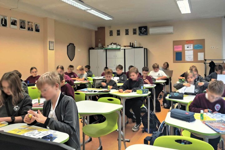Vaizduojama nemaža mokyklinė klasė, kurioje mokiniai susikaupę karpo ir klijuoja įvairias figūras. Klasės priekyje matomi keli tušti suolai. Mokiniai apsirengę tamsiai raudonom arba pilkom uniformom. Klasės gale stovi ant spintelių iškelti istorinių pasta