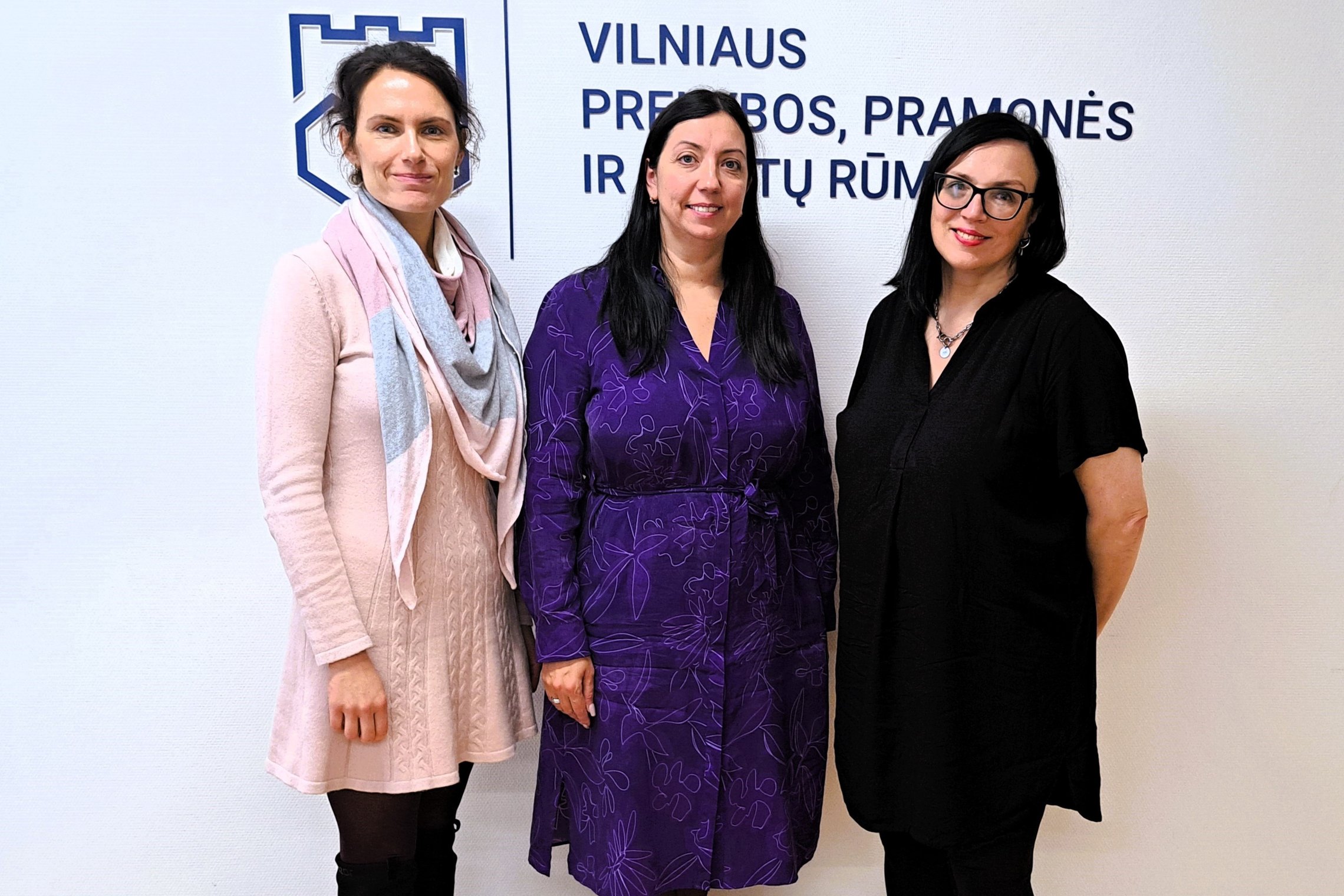 Nuotraukoje trys besišypsančios tamsiaplaukės moterys. Kairėje stovi moteris, vilkinti rausvą suknelę ir pilką šaliką. Vidury moteris su mėlyna suknele, o meteris iš dešinės apsirengusi juodai. Jos draugiškai žvelgia į kamerą. Už nugarų ant sienos matosi 