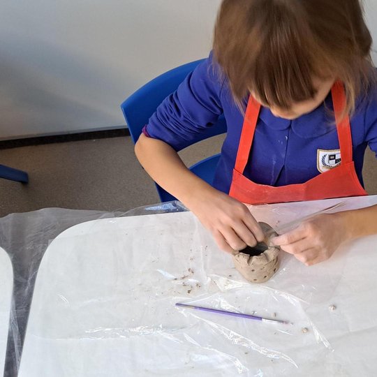 Vaikas, vilkintis mėlynus marškinius ir raudoną prijuostę, sėdi prie stalo ir lipdo molinį indelį. Ant stalo padėtas plastikinis lakštas, molio gabalėliai ir violetinis įrankis lipdymui. Fone matyti knygų lentyna.