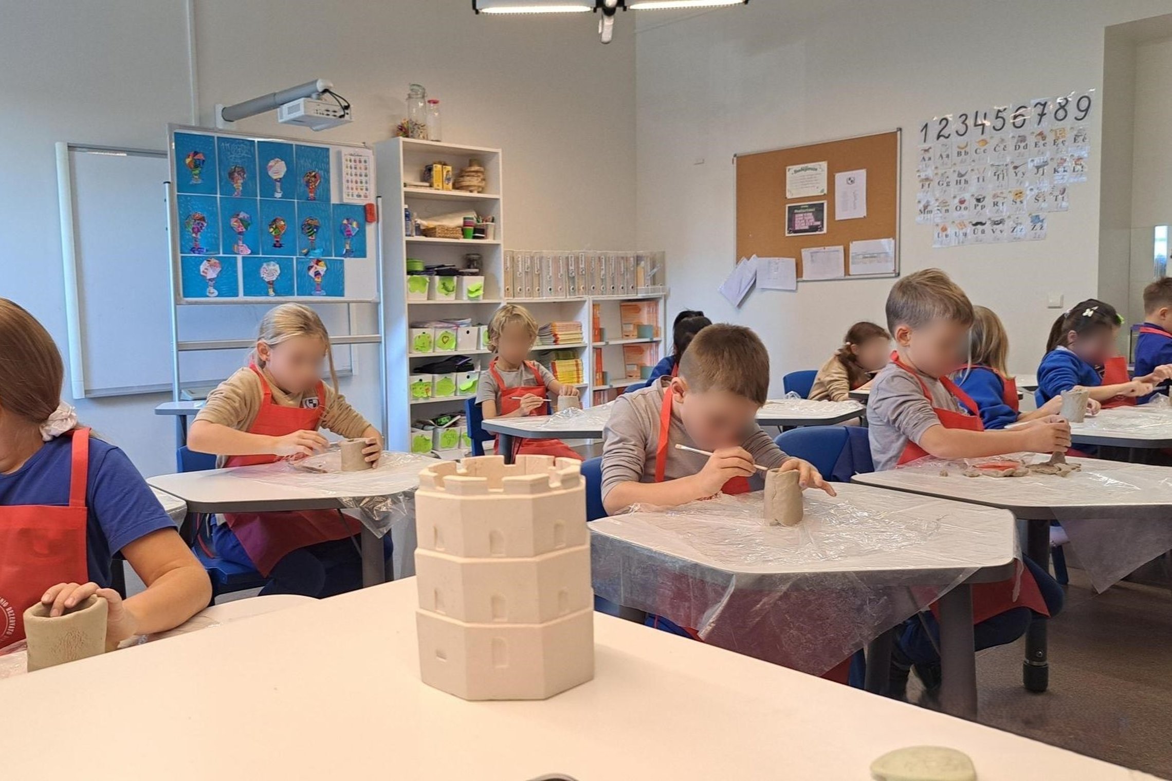 Nuotraukoje pavaizduota klasė pilna vaikų, kurie apsiima keramika. Pirmame plane pavaizduotas Gedimino pilies bokštas, pagamintas iš molio. Visi mokiniai dėvi raudonas prijuostes, ant stalų uždėta plastikinė plievelė, sauganti stalą nuo nešvarumų.