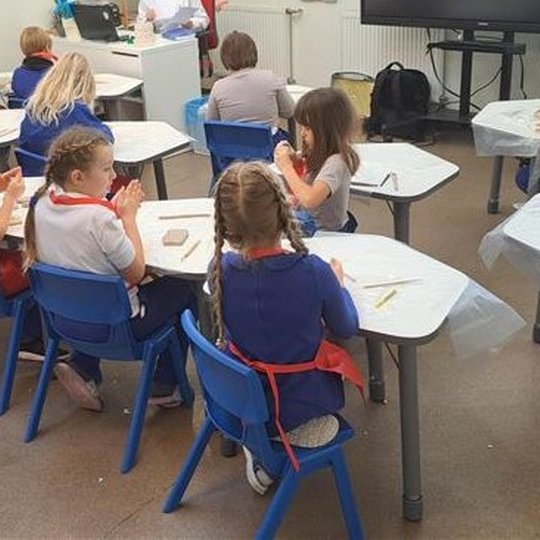 Nuotraukoje matome pradinę klasę, kurioje sėdi pradinukai. Klasė yra atnaujinta, kiekvienas vaikas turi savo stalą ir sėdi ant mėlynų kėdžių. Mokiniai lipdo iš rudojo molio darbelius ir šnekučiuojasi tarpusavyje.
