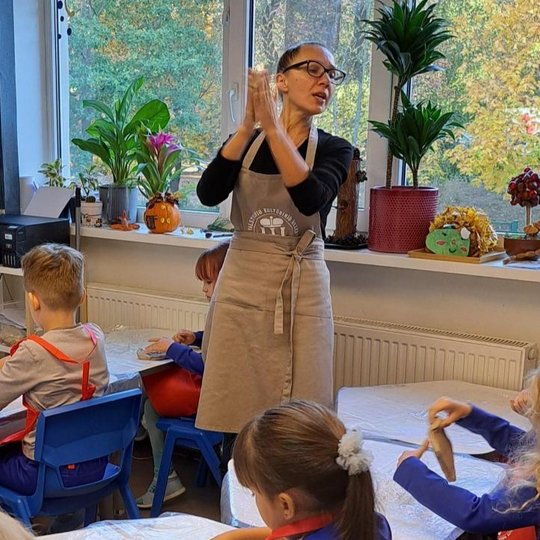 Mokytoja su prijuoste stovi klasėje prie lango ir rodo vaikams, kaip dirbti su moliu, laikydama rankas kartu. Aplink ją sėdi vaikai su raudonomis prijuostėmis, dirbantys su moliu prie stalų. Ant palangės – kambariniai augalai, moliūgų ir rudeninių lapų de