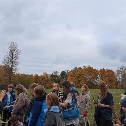 Giedra diena, ant nedidelės kalvos stovi medžiai, kai kurie numetę lapus, kai kurie dar tik rudeniškų spalvų. Matomas būrelis vaikų su mediniais kardais rankose, kurie klausosi gidės pasakojimo. 