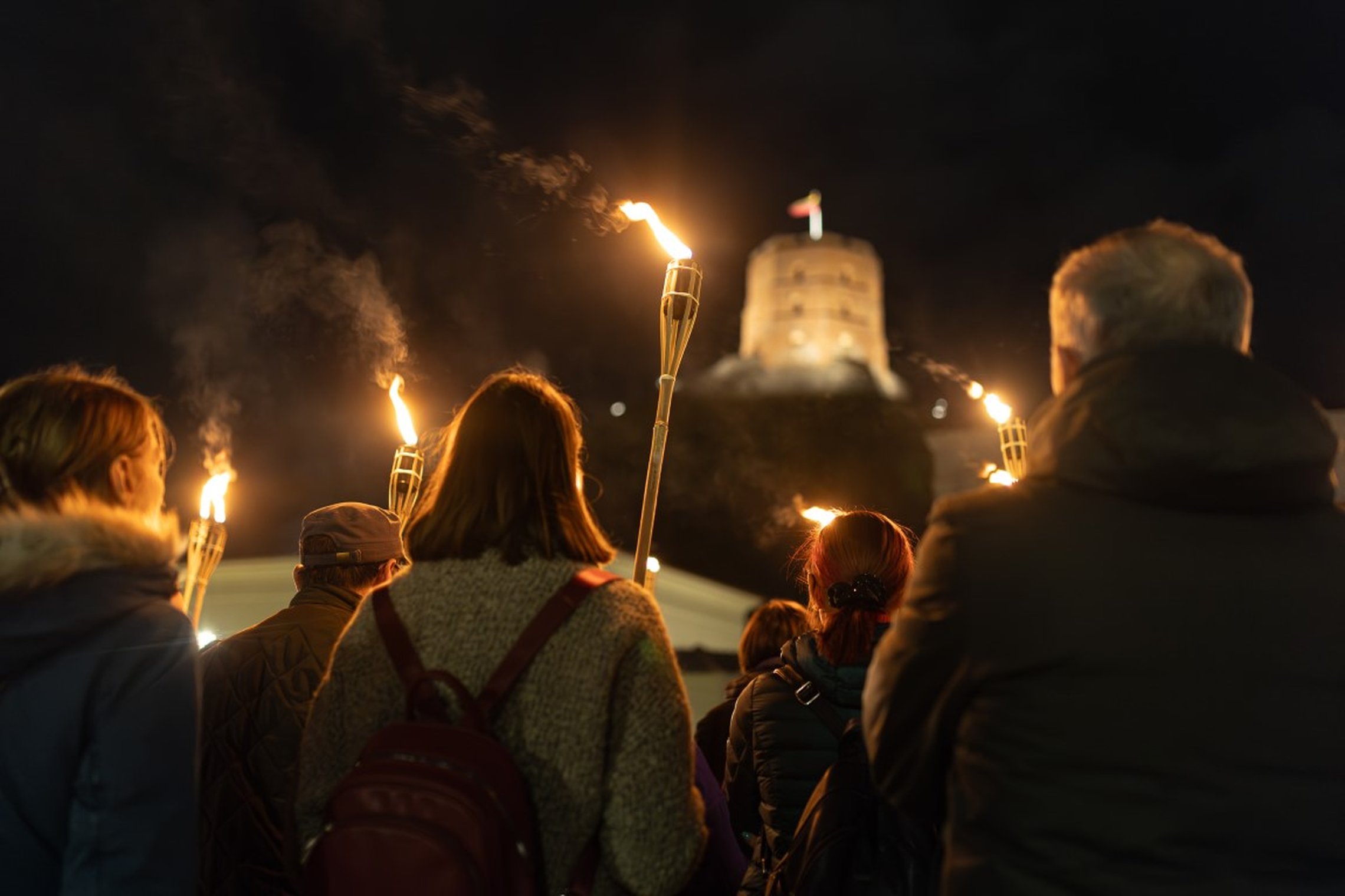 Šiame kadre iš nugaros pusės šiltai apsirengusių žmonių grupė tamsiuoju paros metu. Visi rankose laiko deglus ir žiūri į gražiai apšviestą Gedimino pilies bokšą. Nuotrauka padaryta taip, kad pirmame plane yra žmonės, o pilies bokštas antrame plane.