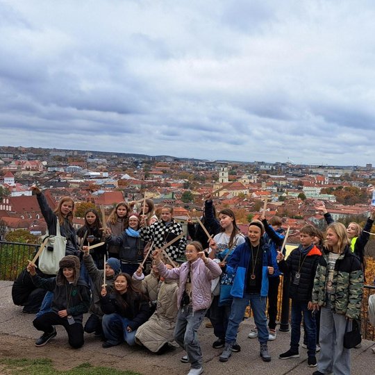 Vaikų būrys pozuoja nuotraukai, visi rankose laiko medinius kardus. Už jų kalno papėdėje matosi senamiesčio pastatų stogai, Gedimino pilis, Valdovų rūmai, rudenio spalvomis pasipuošę medžiai. Vaikai žiūri ne į kamerą, o į žmogų šalia fotografo. 
