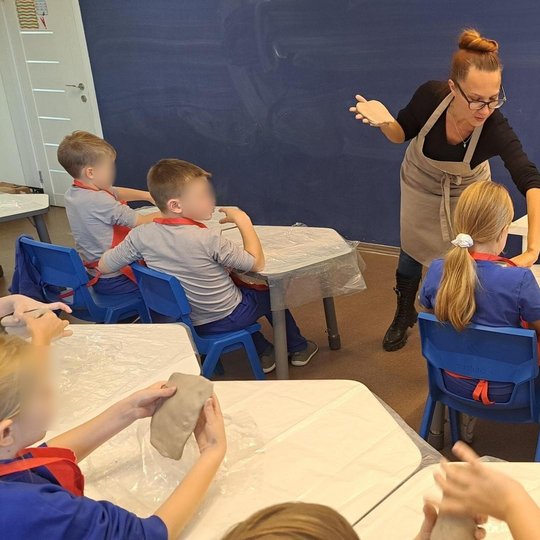 Nuotraukoje pavaizduota klasė su mokinias ir mokytoja. Mokytoja pasilenkus padeda mokiniui su jo keramikos darbu. Visi mokiniai atsisukę nugara į objektyvą. Žiūri į lentą arba į mokytoją ir dirba su moliu.