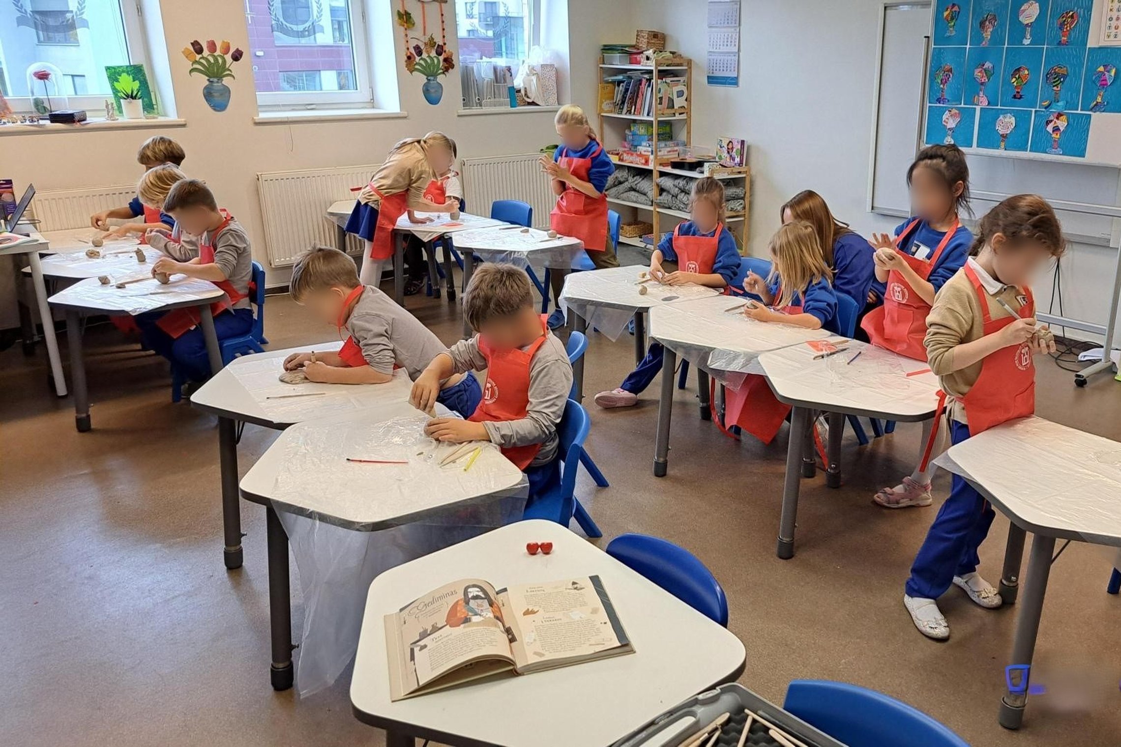 Pradinukai su raudonomis prijuostėmis dirba su moliu klasėje, sėdėdami prie plastikine plėvele dengtų stalų. Klasės priekyje matyti atversta knyga apie kunigaikštį Gediminą, o fone – vaikų darbai, knygų lentyna ir langai su dekoracijomis. Vaikai susikaupę