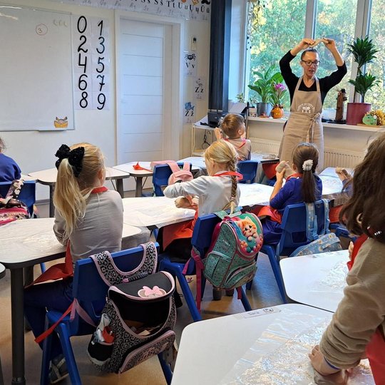 Klasėje vaikai sėdi prie stalų su plastiku dengtais paviršiais, pasiruošę lipdymo veiklai. Priekyje stovi mokytoja su prijuoste, rodanti rankomis judesį, kaip dirbti su moliu. Ant palangės – kambariniai augalai ir rudeniška dekoracija, lange matyti medžia