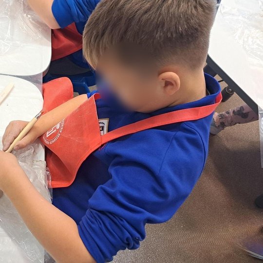 Vaikas su raudona prijuoste, susikaupęs lipdo iš molio, jo rankose yra plastmasinis peiliukas - įrankis padailinti molio dirbinį.
