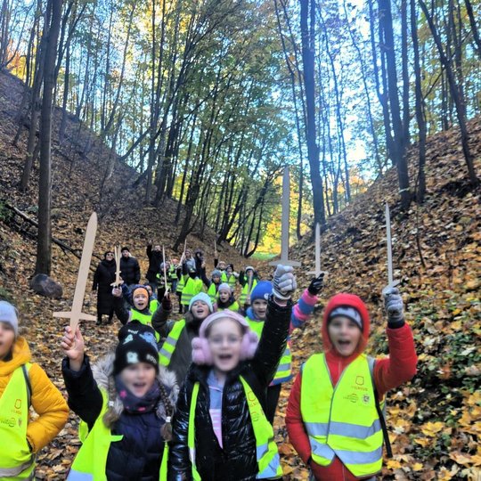 Miške, tarp dviejų kalvų žygiuoja jaunieji keliautojai iškėlę medinius kalavijus į viršų. Jie ,apsirengę ryškiomis, šviesą atspindinčiomis liemenėmis, žygiuoja per rudens lapų nusėtą mišką ir atrodo tokie smulkūs palyginus su miško mastu.