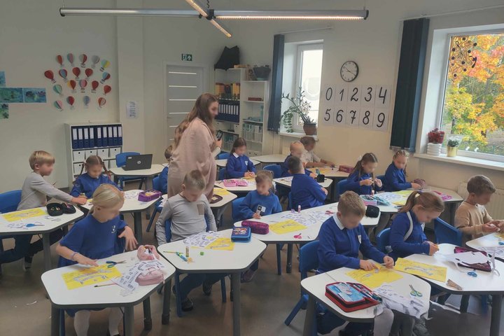 Vaizduojama pradinės mokyklos amžiaus mokinių klasė. Vaikai susikaupę karpo ir klijuoja įvairias figūras. Dauguma jų dėvi mėlynas uniformas. Vaikų darbą stebi tarp suolų einanti mokytoja apsirengusi rausvą megztinį. Nuotrauka daryta rudenį, nes pro langą