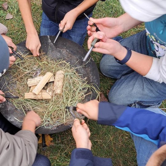Nuotrauka: penki vaikai bando užkurti laužą į metalinę lazdelę trindami kitą metalinę lazdelę laužinėje sudėję sudžiūvusią žolę ir ant viršaus daug smalėkų. Veiksmas vyksta lauke. Vaikai apsirengę lauko rūbais ir aplinkui yra žolė. 