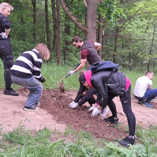 Grupė žmonių dirba miško aikštelėje – kasa žemę, lygina paviršių ir formuoja taką. Kai kurie naudoja kastuvus, kiti rankomis tvarko smėlį ir žemes. Aplink – tanki žaluma, medžiai, pavasariška gamta. Dalyviai dėvi pirštines ir patogią lauko aprangą, atmosf