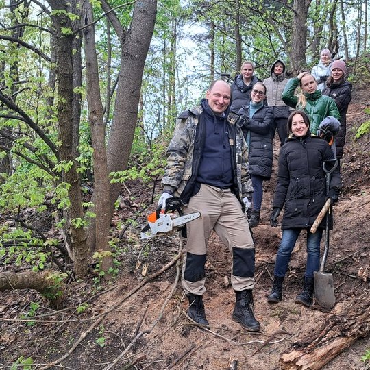 Grupė žmonių stovi miško šlaite, apsirengę šiltais lauko drabužiais. Priekyje vyras laiko motorinį pjūklą, moteris – kastuvą. Aplink matyti nupjauti medžiai, šakos ir takas per mišką. Visi šypsosi, atmosfera pozityvi – akivaizdu, kad dalyvauja bendroje mi