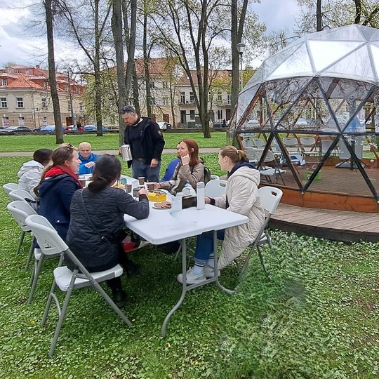 Grupė žmonių sėdi aplink stalą parke, geria arbatą ir bendrauja. Stalas pastatytas ant žolės šalia kupolo formos skaidraus paviljono, pažymėto Vilniaus pilių ženklu. Aplink matyti miesto pastatai, medžiai ir pavasarinė žaluma. Vaizdas perteikia bendruomen