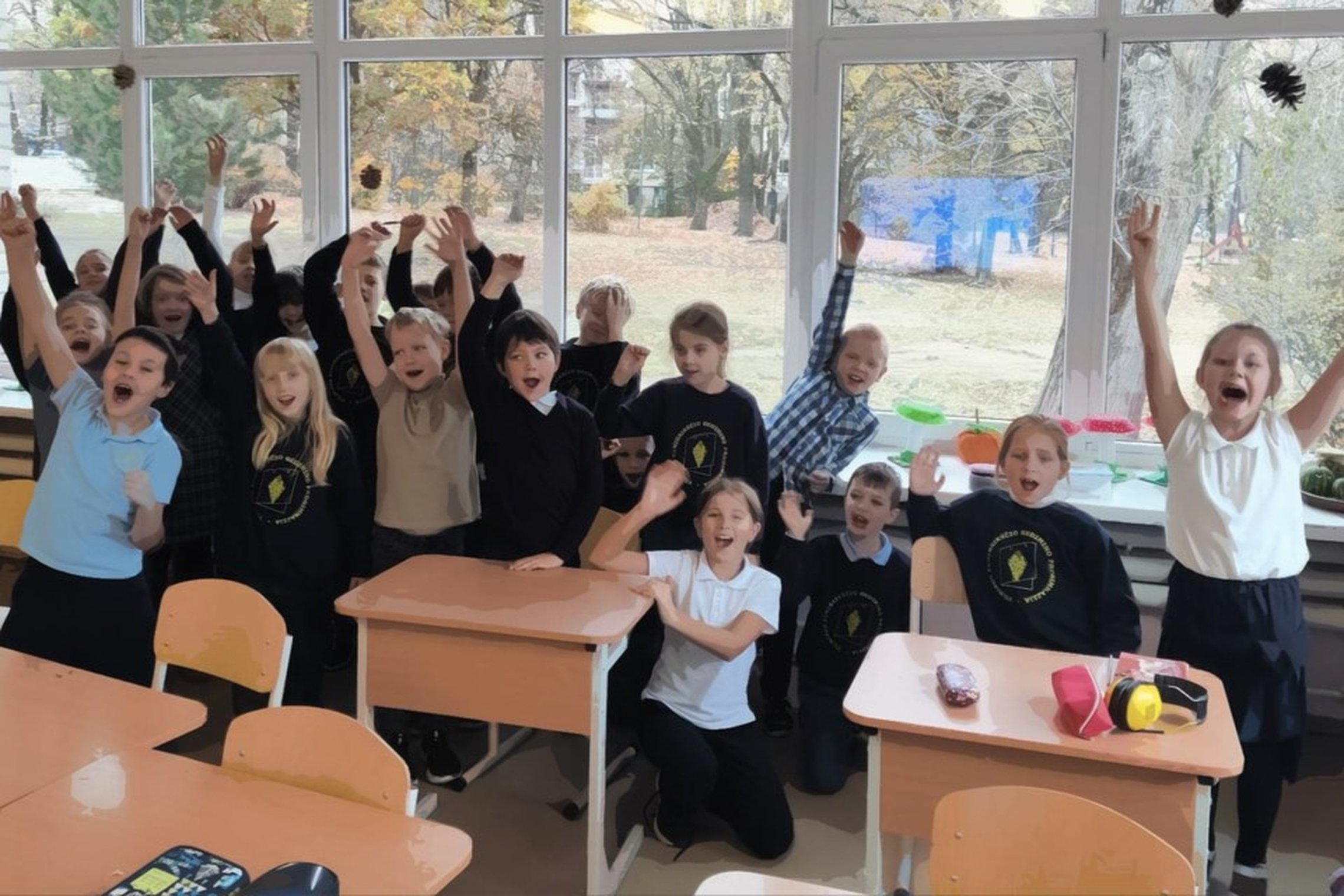 Džiugiai nusiteikusi vaikų grupė klasėje stovi ar sėdi prie suolų, daugelis pakėlę rankas į viršų. Vaikai vilki uniformas su mokyklos logotipu arba margus marškinėlius. Už jų – dideli langai, pro kuriuos matyti rudeninis peizažas su medžiais ir nukritusia