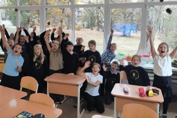 Džiugiai nusiteikusi vaikų grupė klasėje stovi ar sėdi prie suolų, daugelis pakėlę rankas į viršų. Vaikai vilki uniformas su mokyklos logotipu arba margus marškinėlius. Už jų – dideli langai, pro kuriuos matyti rudeninis peizažas su medžiais ir nukritusia