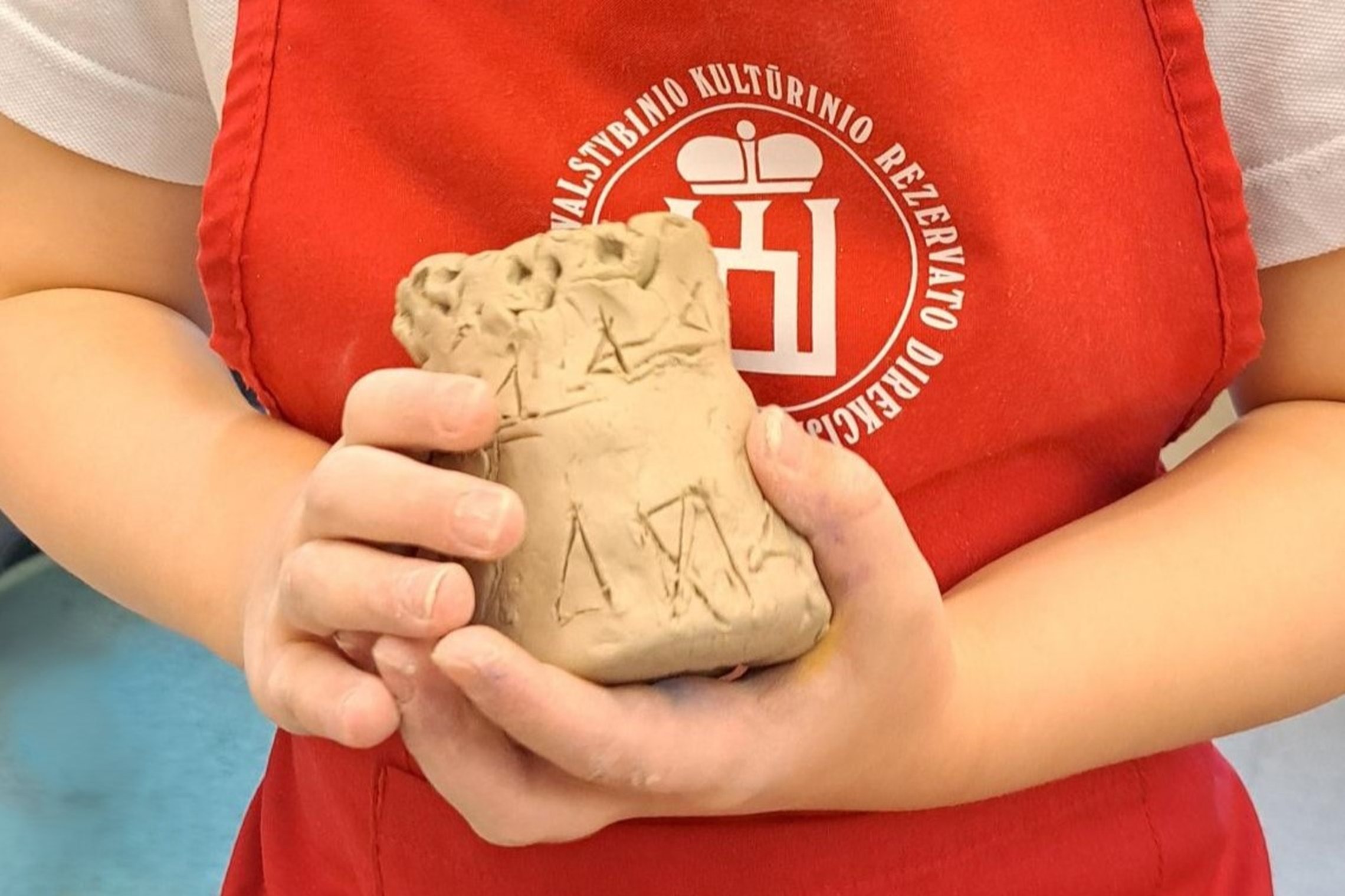 Nuotraukoje matomas lipdinys iš molio ant kurio yra daug ornamentų. Šį lipdinį laiko vaikas, kuris dėvi raudona su Vilniaus pilių emblema prijuoste.