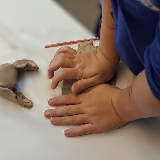 Vaiko rankos spaudžia molį ant plastiku dengto stalo, šalia – jau suformuota molio figūrėlė, primenanti gyvūną ar fantastinį padarą. Ant stalo taip pat guli medinis lipdymo įrankis ir raudonas pieštukas. Vaikas vilki mėlynus marškinėlius.
