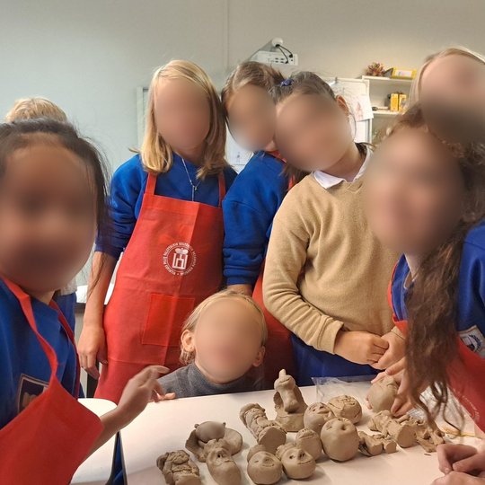 Nuotraukoje vaizduojamos septynios mergaites sustojusios aplink stalą, jų veidai paslėpti. Ant stalo sudėta daugybė molinių galvų su burnomis, nosimis, akimis.