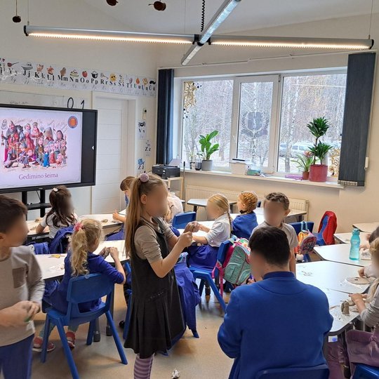 Pradinukų klasė, kurioje vyksta edukacinė veikla apie Gedimino šeimą – priekyje rodomas skaidrių ekranas su spalvinga iliustracija ir užrašu „Gedimino šeima“. Vaikai sėdi prie stalų ir lipdo iš molio, kai kurie stovi ir dirba. Klasėje šviesu, ant palangių