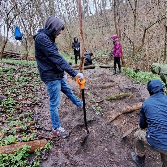 Keletas žmonių su striukėmis ir gobtuvais dirba miško šlaite lietingą dieną. Jie kasa, tvarko ir įrenginėja taką iš medžių kamienų bei žemių. Aplinka šlapia, purvina, gausiai apaugusi žema augmenija ir nuogais medžiais. Akivaizdu, kad vyksta savanoriška m
