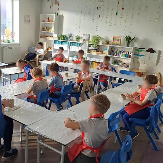 Nuotraukoje matome klasėje sėdinčių trylika vaikų ir vieną auklėtoją. Dvylika vaikų, kartu su auklėtoja, lipdo iš molio Gedimino bokštą. Klasės kampe krėsle sėdi vaikas pasidėjęs nešiojimą kompiuterį ant kelių. Priešais vaikus ant stalo yra pastatytas kom