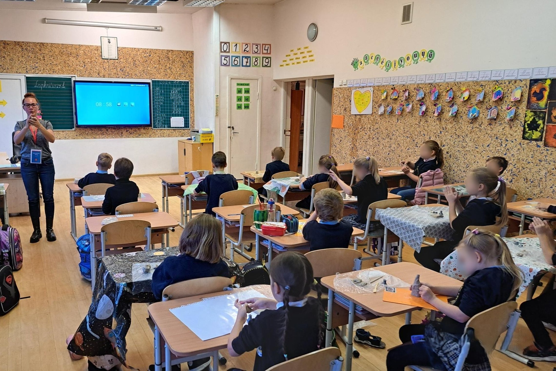 Nuotraukoje vaizduojama kalsė, fotografuota iš galo. Klasės priekyje išmanioji lenta rodo laiką, sienos papuoštos mokomosiomis iliustracijomis: skaičiais raidėmis. Prieš lentą stovi edukatorė ir vaikams kažką aiškina, vaikai sėdi suoluose po vieną, ant st