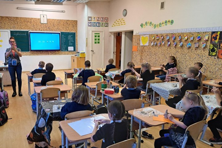 Nuotraukoje vaizduojama kalsė, fotografuota iš galo. Klasės priekyje išmanioji lenta rodo laiką, sienos papuoštos mokomosiomis iliustracijomis: skaičiais raidėmis. Prieš lentą stovi edukatorė ir vaikams kažką aiškina, vaikai sėdi suoluose po vieną, ant st