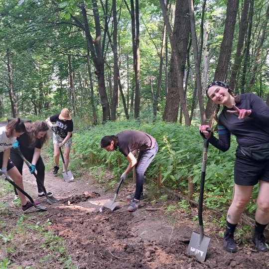 Šešios moterys miško take kasa žemę kastuvais, tvarko ir lygina paviršių. Aplinka – žaluma, aukšti medžiai, vasariška gamta. Viena moteris pozuoja su šypsena, rodo taikos ženklą, kitos susitelkusios į darbą. Vaizdas atspindi aktyvią savanorystę ar bendruo