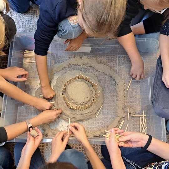 Vaizduojami vaikai prie plastikinės dėžės, kurioje puikuojasi jų pastatytas piliakalnis. Mokiniai kruopščiai išlipdė piliakalnio kontūrus, mediniais pagaliukais imitavo tvorą, iš kartono pastatė nedidelę pilį ir tiltą, vedantį į ją.